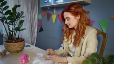 Güzel kızıl saçlı bir kız, yün sarıp oyuncak yapıyor. Kıza yün keçeli iğne batırılmış. El yapımı iş