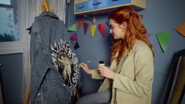 Ein schönes rothaariges Mädchen malt mit bunten Farben auf Kleidung. Handarbeit — Stockvideo