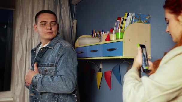 Ένας νεαρός άνδρας δοκιμάζει ένα τζιν σακάκι με ένα ζωγραφισμένο αποτύπωμα. Μια κοπέλα βγάζει φωτογραφία έναν τύπο με μπουφάν στο κινητό της. Χειροποίητη εργασία — Αρχείο Βίντεο