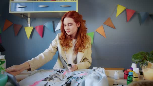 Obecný plán krásné rusovlasé dívky kreslit s pestrobarevnými barvami na oblečení. Dívka se dívá na video na svém telefonu a kreslí. Ruční práce — Stock video
