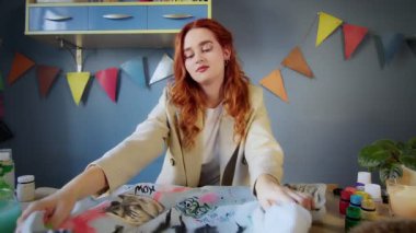 Güzel, kızıl saçlı bir kız elbiselerine renkli boyalar çiziyor. Kız bitirilmiş eserlere bakıyor. El yapımı iş