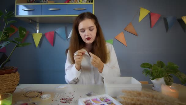 Una hermosa chica con una camisa blanca crea una decoración de cuentas. Trabajo hecho a mano — Vídeo de stock