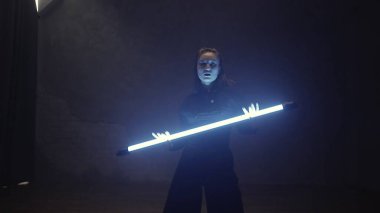 A beautiful girl is dancing indoors in a black suit with a blue lamp in her hands. Dances