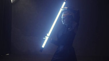 A beautiful girl is dancing indoors in a black suit with a blue lamp in her hands. Dances