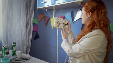 Güzel kızıl saçlı bir kız çantasına rengarenk boyalar çiziyor. Kız bitirilmiş eserlere bakıyor. El yapımı iş