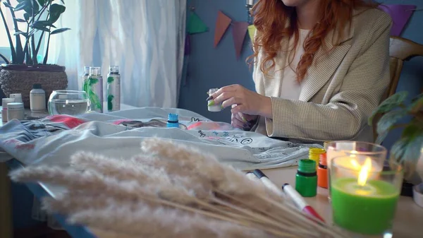 Close-up of a beautiful red-haired girl drawing with multicolored paints on clothes. Handmade work — Stockfoto