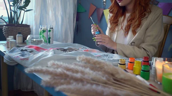 Close-up of a beautiful red-haired girl drawing with multicolored paints on clothes. Handmade work — Stock Photo, Image