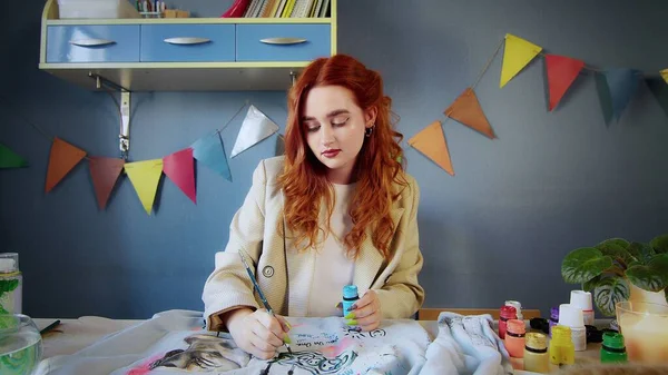 El plan general de una hermosa chica pelirroja dibujando con pinturas multicolores en la ropa. Trabajo hecho a mano —  Fotos de Stock