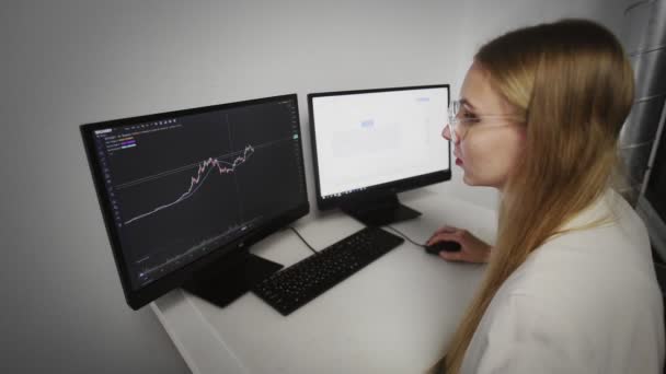 La ragazza è seduta al computer e guarda il grafico del mercato azionario online che mostra le tendenze orecchiabili e rialziste della valuta Bitcoin. In tempo reale — Video Stock