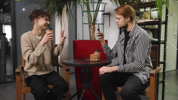 Dos gays en una cita están sentados en una cafetería, bebiendo café y tomando fotos el uno del otro por teléfono. LGTB — Vídeo de stock