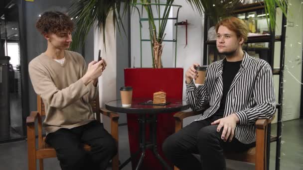 Δύο γκέι άντρες σε ραντεβού κάθονται σε μια καφετέρια, πίνουν καφέ και βγάζουν φωτογραφίες ο ένας τον άλλον στο τηλέφωνο. ΛΟΑΤ — Αρχείο Βίντεο
