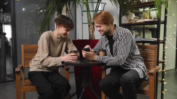 Δύο γκέι άντρες σε ραντεβού κάθονται σε ένα καφέ, πίνουν καφέ και κοιτάζουν φωτογραφίες στο κινητό τους. ΛΟΑΤ — Αρχείο Βίντεο
