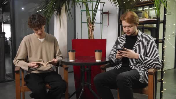 Dois gays num encontro estão sentados num café. Um tipo está a ler um livro e o outro está a escrever uma mensagem no telemóvel. LGBT — Vídeo de Stock