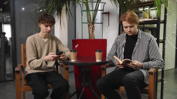 Dos gays en una cita están sentados en una cafetería. Un tipo está leyendo un libro y el otro está escribiendo un mensaje en su teléfono. LGTB — Vídeo de stock