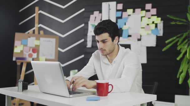 Een blanke jongeman zit achter een computer en werkt op afstand, maakt aantekeningen op papier. Een jongeman die op afstand werkt. — Stockvideo