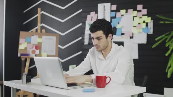 En ung mand af kaukasisk udseende sidder ved en computer og arbejder eksternt og laver noter på papir. En ung person, der arbejder eksternt, drikker kaffe eller te – Stock-video