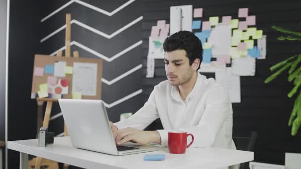 Un joven cansado de apariencia caucásica está sentado en una computadora y trabajando remotamente. Un joven trabajando remotamente — Vídeos de Stock