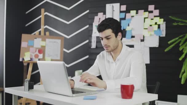 Счастливый молодой человек кавказской внешности сидит за компьютером и работает удаленно. Молодой человек, заканчивает работу — стоковое видео