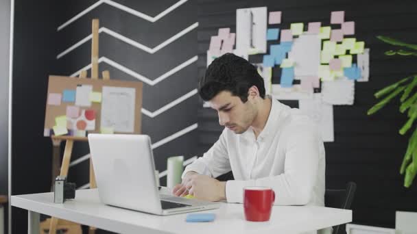 Um jovem de aparência caucasiana está sentado em um computador e trabalhando remotamente, fazendo anotações em papel. Um jovem trabalhando remotamente — Vídeo de Stock