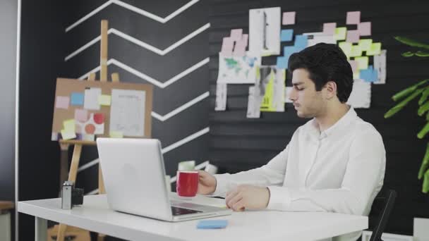 Молодой человек кавказской внешности сидит за компьютером, глядя в окно и пьет чай или кофе. Молодой человек работает дистанционно — стоковое видео