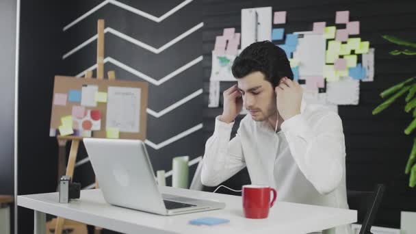코카서스 출신의 젊은 남자가 컴퓨터 앞에 앉아 원격으로 일하고 있습니다. 젊은 남자가 헤드폰을 끼고 비디오 링크를 통해 의사소통을 한다 — 비디오