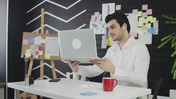 Молодой кавказский человек сидит за компьютером и работает удаленно. Молодой человек в наушниках общается по видеосвязи и показывает свои заметки на стене — стоковое видео