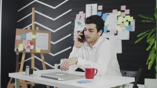 Молодой кавказский мужчина сидит за столом и разговаривает по телефону. Молодой человек работает дистанционно — стоковое видео