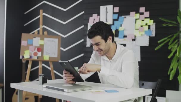 Un joven de apariencia caucásica está sentado en una mesa en una tableta y sitios web de navegación y. Un joven trabajando remotamente — Vídeo de stock