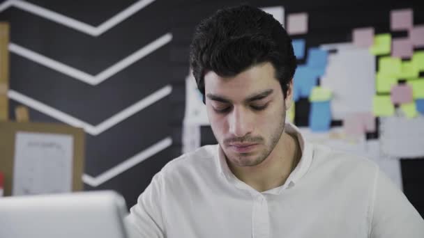 Close-up af en ung mand af kaukasisk udseende sidder ved en computer og arbejder eksternt, gøre noter på papir. En ung mand, der arbejder på afstand. Video i bevægelse – Stock-video