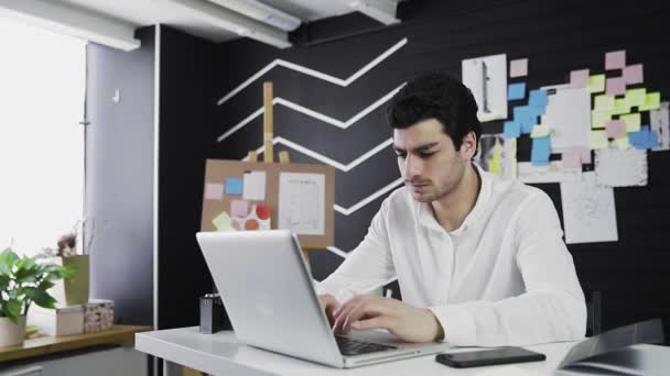 En ung mand af kaukasisk udseende sidder ved en computer og arbejder på afstand. Den unge mand tager telefonen og tjekker den. Video i bevægelse – Stock-video