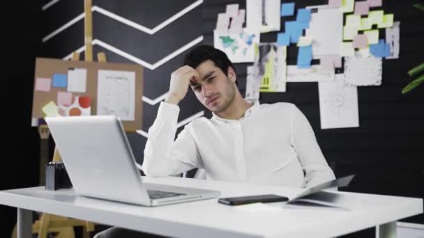 Задумчивый молодой человек с кавказской внешностью сидит за компьютером. Молодой человек работает дистанционно. Видео в движении. Фрилансер. Приближается к камере — стоковое видео