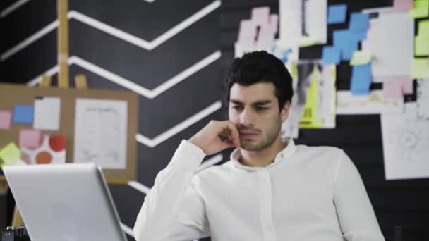 Задумчивый молодой человек с кавказской внешностью сидит за компьютером и смотрит в сторону от компьютера. Молодой человек работает дистанционно. Приближается к камере — стоковое видео