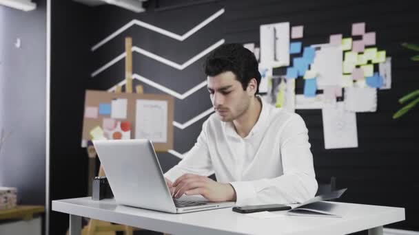 En ung mand af kaukasisk udseende sidder ved en computer. Fjernarbejde. Freelance. Zoomer ind på kameraet – Stock-video