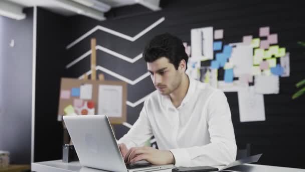 Um jovem de aparência caucasiana está sentado em um computador, olhando para a câmera. Trabalho remoto. Freelance. A fazer zoom na câmara — Vídeo de Stock