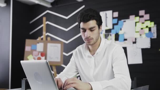 Un tânăr de înfățișare caucaziană stă la un computer, se uită furios la cameră. Muncă de la distanţă. Freelance. Distanța camerei — Videoclip de stoc
