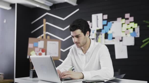 Un joven de apariencia caucásica está sentado frente a una computadora, sonriendo y mirando a la cámara. Trabajo remoto. Independiente. Acercamiento de la cámara — Vídeo de stock