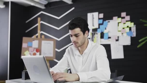 En ung mand af kaukasisk udseende sidder ved en computer og ser vredt på kameraet. Fjernarbejde. Freelance. Zoomer ind på kameraet – Stock-video