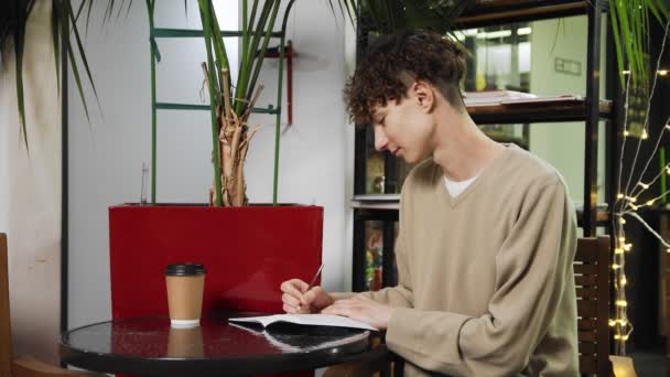 Primer plano de un tipo sentado en un café, bebiendo café y escribiendo en un cuaderno. Trabajar fuera de casa — Vídeo de stock