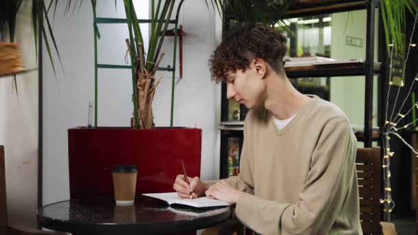 Close-up de um cara sentado em um café e escrevendo em um caderno. A empregada traz-lhe café. Cafeteira — Vídeo de Stock