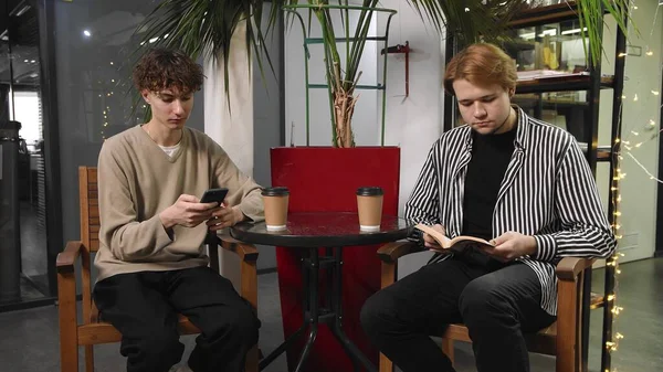 Two gay guys on a date are sitting in a coffee shop. One guy is reading a book and the other is typing a message on his phone. LGBT — Stock Photo, Image