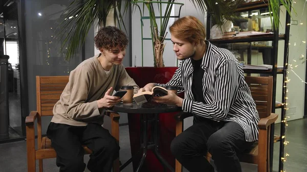 Two gay guys on a date are sitting in a coffee shop. One guy is reading a book and the other is typing a message on his phone. LGBT — Stock Photo, Image