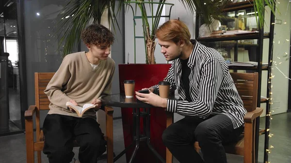 Dos gays en una cita están sentados en un café. Un tipo está leyendo un libro y el otro está mostrando algo en su teléfono. LGTB — Foto de Stock