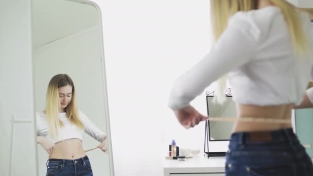 Uma jovem está na frente de um espelho e mede a circunferência da cintura com uma fita métrica e está feliz com os indicadores. — Vídeo de Stock