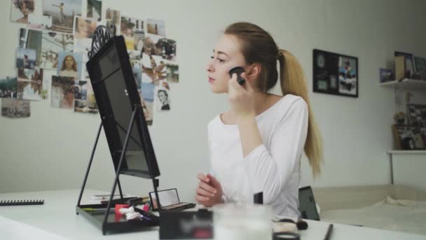 Uma jovem faz sua maquiagem enquanto se senta em uma mesa na frente de um espelho — Vídeo de Stock