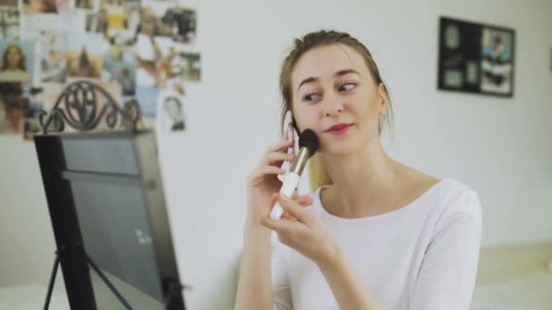 Une jeune femme utilise un pinceau de maquillage pour dessiner un fard à joues. La fille parle au téléphone et fait son maquillage — Video