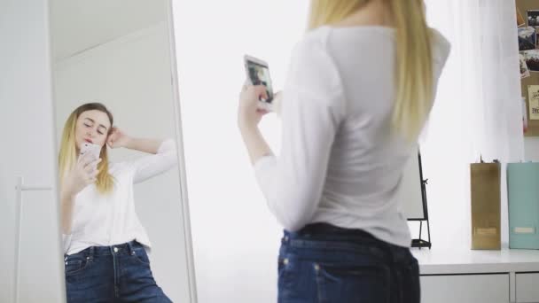 A young woman looks in the mirror, preens herself, takes pictures of herself on her phone in the mirror, adjusts her clothes and hairstyle. He smiles and likes himself. — Stock Video