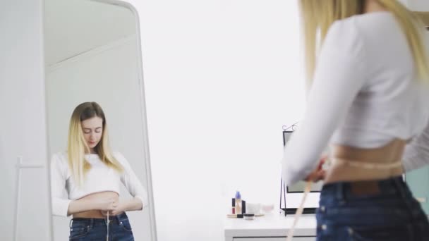 Uma jovem mulher fica na frente de um espelho e mede a circunferência da cintura com uma fita métrica. A menina é complexa por causa do peso — Vídeo de Stock