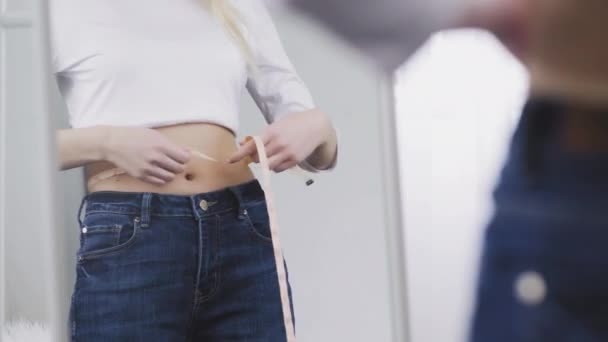 Uma jovem mulher fica na frente de um espelho e mede a circunferência da cintura com uma fita métrica — Vídeo de Stock