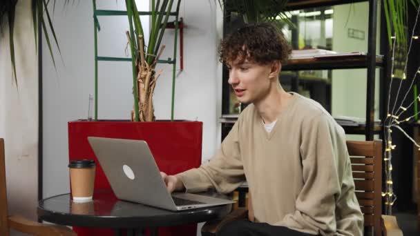 Primer plano de un tipo sentado en un café con una computadora portátil en una mesa, bebiendo café y hablando a través de un enlace de vídeo. Trabajar fuera de casa — Vídeo de stock