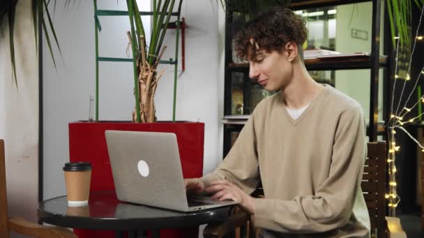 Primer plano de un tipo sentado en una cafetería con un portátil en la mesa, bebiendo café y escribiendo en el teclado. Trabajar fuera de casa — Vídeo de stock